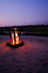 Metal brazier burning on a sandy beach at dusk by the Okavango River, with soft twilight colors and footprints in the sand.