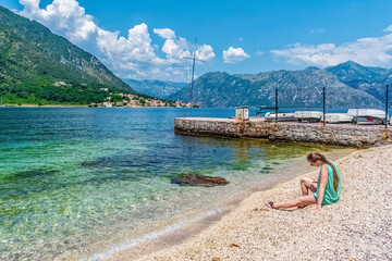 A peaceful scene at a beach in Kotor, Montenegro, with a woman sitting on the pebbled shore. The stunning blue waters and surrounding mountains create a picturesque and relaxing atmosphere