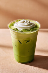 Iced matcha latte with soft cream topping in clear plastic cup on beige surface, refreshing and creamy drink