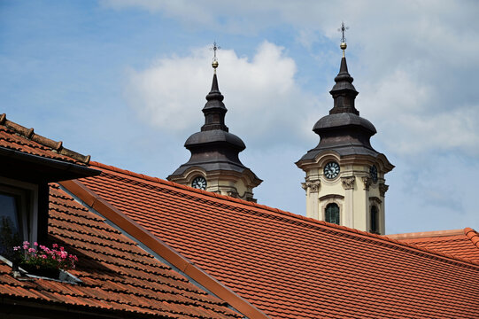 catholic church clock and bell towers in a distance with white clock faces. Baroque style architecture. brown clay tile sloped house roof in the foreground. dormer and window. pink flowers. tourism. - Powered by Adobe