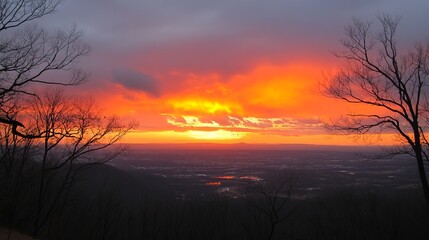 Fototapeta premium Fiery sunset over a valley, viewed from a mountaintop through bare, silhouetted trees. The sky blazes with intense oranges and reds