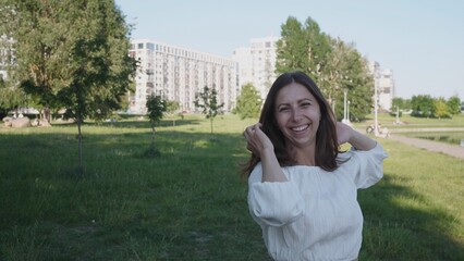 A cheerful woman stands in a lush park, playfully touching her hair while showcasing her smile against a backdrop of urban architecture and greenery.