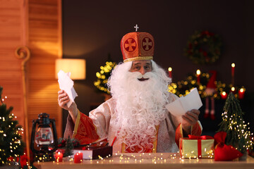 Saint Nicholas with letters at desk in room decorated for Christmas