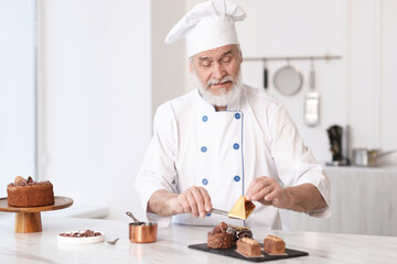 Elderly pastry chef decorating tasty desserts with edible gold leaf sheet at table in kitchen