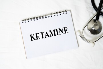 The medical concept "KETAMINE" is written clearly on a white spiral notebook, accompanied by a stethoscope on a white background.