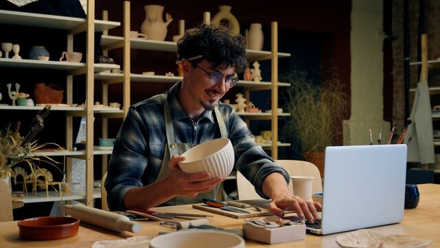 Male potter artist Caucasian guy man small business owner salesman seller designer sculptor typing laptop online selling ceramics store commerce handmade crafting studio art decorator pottery workshop
