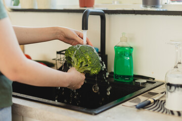 Woman washing fresh broccoli under running water in kitchen sink. Healthy eating and food hygiene concept.
