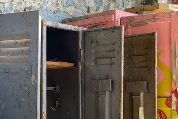 Damaged metal lockers with open doors, placed against a peeling wall in an abandoned building. Hooks and shelves are visible inside. The scene has an industrial, post-apocalyptic atmosphere.