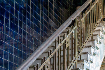 Fototapeta premium Damaged concrete staircase with a decorative metal railing, running along a wall covered with dark blue tiles. The scene captures a neglected piece of industrial interior architecture.