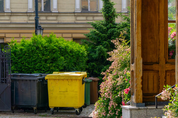 Waste sorting bins placed near a fence in an urban courtyard, surrounded by dense greenery and...