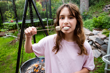 A 12-year-old brown-eyed girl toasting marshmallows on charcoal grill in backyard. A cozy moment of...