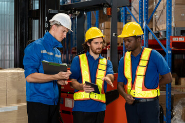 Three male warehouse workers discussing logistics tasks using digital tablet and clipboard while wearing safety helmets and reflective vests in storage facility surrounded by racks and cardboard boxes