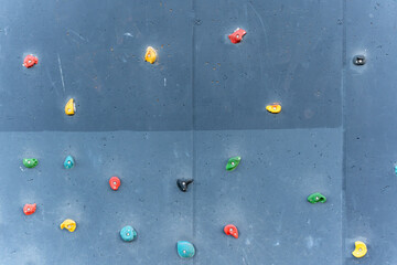 A vertical climbing wall with colorful holds against a gray background. The various shapes and colors offer a playful, challenging design for sport or playground use.