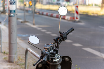 A scooter is parked on the side of the road, the focus is on the handlebars with mirrors. The road and red and white bollards are visible in the background.