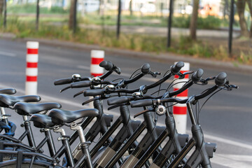 A row of black rental bicycles stands neatly next to each other, with red and white bollards in the background. The photo shows a municipal bike rental system.