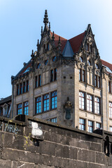 An elaborately designed historic building with red roofs and pointed gables rises into the blue sky. The facade is richly decorated with ornaments and coats of arms.