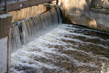 A small waterfall flows over a weir into a river. The structure demonstrates the combination of industrial construction and flowing water.