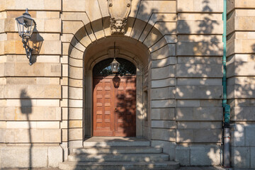 An old stone entrance portal with a massive wooden door and a classic lantern. Light and shadow emphasize the details of the sandstone facade.