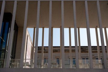An open terrace with a glass railing and many empty chairs, framed by vertical concrete columns. Classic buildings can be seen in the background.