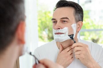 Handsome man shaving with razor near mirror at home
