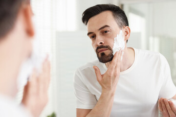 Handsome man applying shaving foam near mirror in bathroom