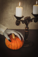 Vintage candelabrum with burning candles and pale vampire hand with touches small pumpkin on the table, vertical image.