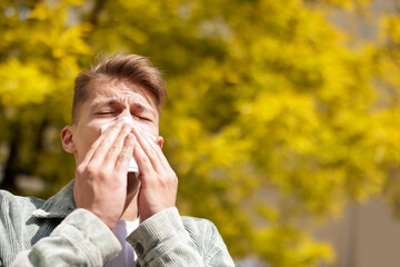 Young man with paper tissue suffering from seasonal pollen allergy near tree on spring day. Space...
