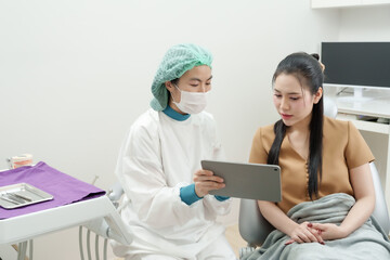 asian male dentist wearing surgical cap mask gown using digital tablet to explain root canal...