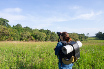 An Asian woman uses binoculars to explore the forest, carrying a backpack equipped with a solar panel charger, symbolizing sustainable hiking, off-grid adventure, and eco-friendly travel.