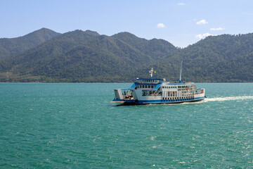 Passing ferry on azure waters near Koh Chang island, creating white wake under sunny skies with forested hills in background. Thailand.

