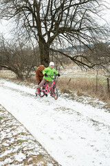 Obraz premium Winter fun unfolds as a child rides a pink bicycle while being pushed through a snowy path by an adult in a rural landscape surrounded by bare trees and frosty fields
