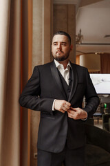A man with a beard puts on a jacket by the window.