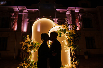 Silhouettes of bride and groom in front of a wedding arch decorated with flowers