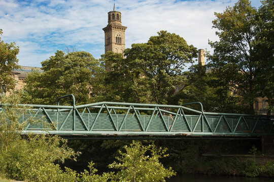 The metal bridge spanning the River Aire that links the world heritage site village of Saltaire with Roberts Park with the chimneys of Salts Mill in the background