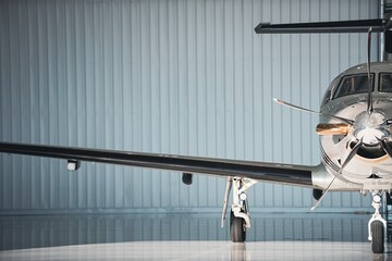 Front partial view of a stationary turboprop aircraft in a hangar, framed off-center with landing gear and propeller visible under soft directional light. © Paul
