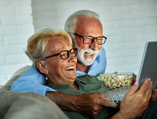 Portrait of an elderly senior happy couple using laptop at night, using modern technology having fun with glowing screen in dark office or at home, watching tv together eating popcorn at home. Shot of
