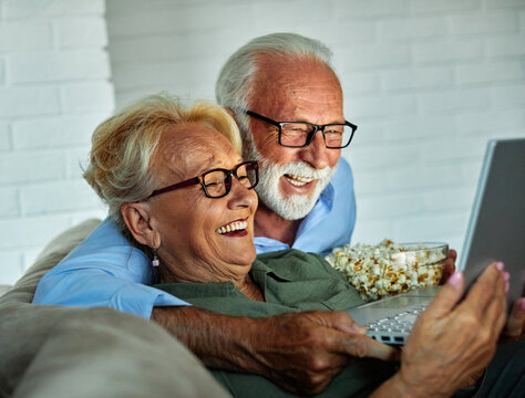 Portrait of an elderly senior happy couple using laptop at night, using modern technology having fun with glowing screen in dark office or at home, watching tv together eating popcorn at home. Shot of