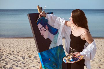 Young female artist in black dress paints portrait on canvas set against serene seaside. Scene...