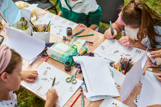 Young hands sketching at table filled with art supplies in sunny outdoor setting. Green grass visible, vibrant atmosphere enhances creativity and focus