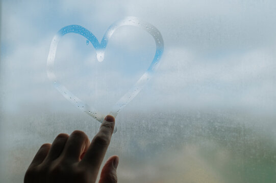 A hand drawing a heart on a foggy window during a rainy day