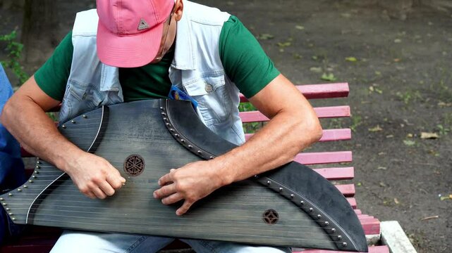 A musician in a pink cap plays a vintage wooden gusli while sitting on a bench in the park