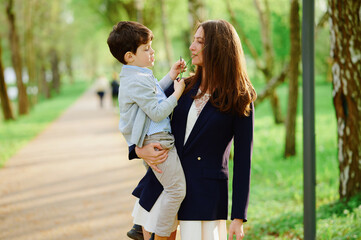 mother and son bond outdoors in springtime park setting. woman in blazer holds young boy close on...