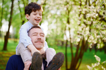father carrying young son on shoulders in lush spring park, sharing joyful laughter. scenic nature background enhances family bonding moment, creating heartwarming outdoor experience