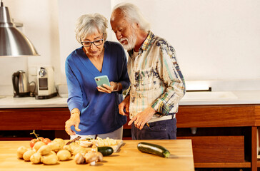 Senior couple using smartphone and preparing vegetables in kitchen