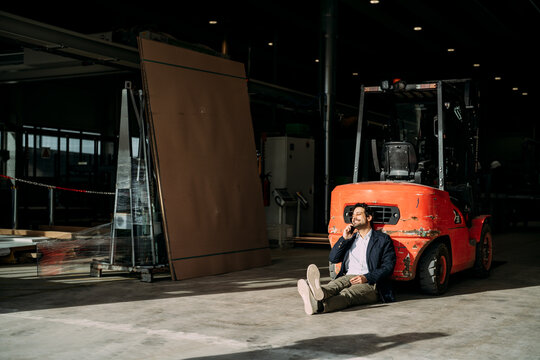 Man sitting on the floor in a warehouse talking on the phone near a forklift