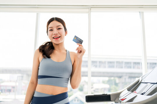 Smiling young woman in active wear holding a credit card while standing near a treadmill in a modern gym. Concept of healthy lifestyle, fitness membership, digital payment, and online shopping.
