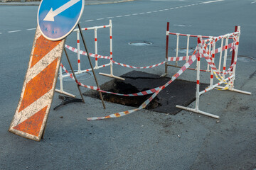 Blue arrow pointing to the left is on a sign. The sign is next to a road with a hole in it. There...
