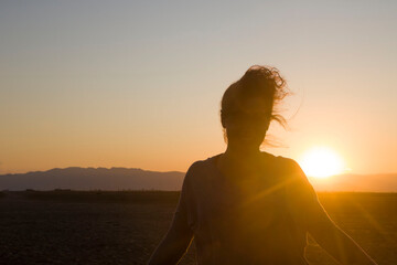 Young woman on the beach at sunset. The sun sets against the sand and trees, and the woman in the...