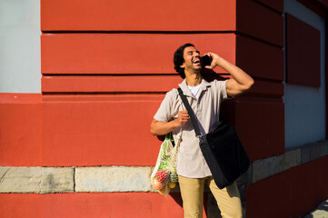 Happy indian businessman talking on smartphone while carrying groceries in a reusable bag,...