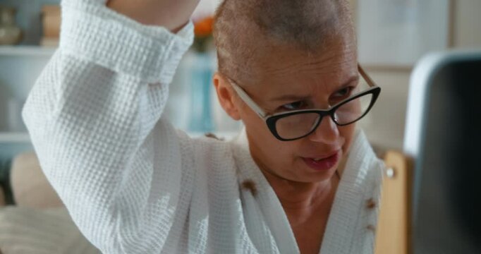 Determined and confident adult woman with cancer, after chemotherapy, is cutting her hair, shaving her head with a hair clipper at home in the living room.